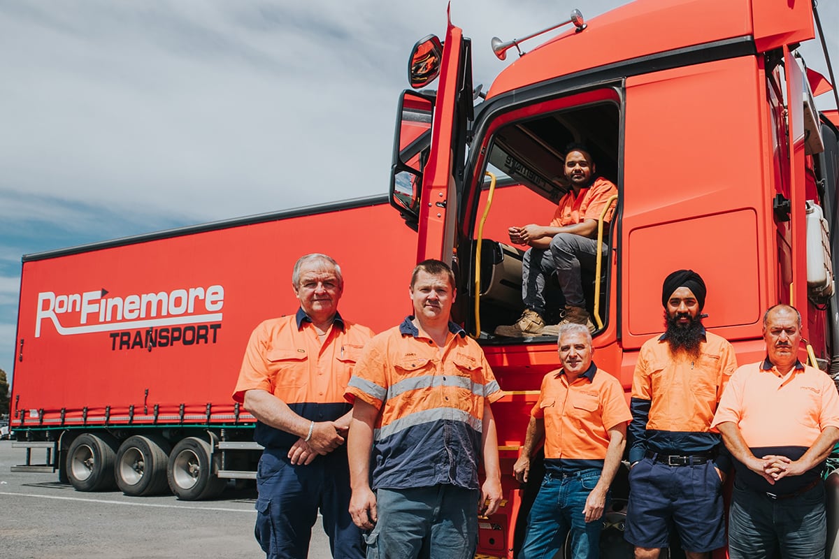 Ron Finemore Transport employees standing next to a red truck, wearing orange high-visibility uniforms. A man sits inside the truck, while others stand outside, representing the diverse team.