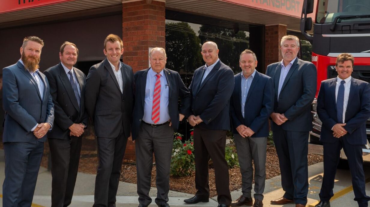 A group of Ron Finemore Transport executives in formal attire, standing in front of the company’s office building with the logo visible above.